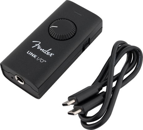 The image shows a matte black Fender Link I/O audio interface, a compact digital device for music recording. It features a large volume knob, a 3.5mm headphone jack, and a USB-C connection for computers or mobile devices. Designed for portability, it enables musicians to record instruments, monitor audio with low latency, and convert analog signals to digital, ideal for guitarists and home studios.