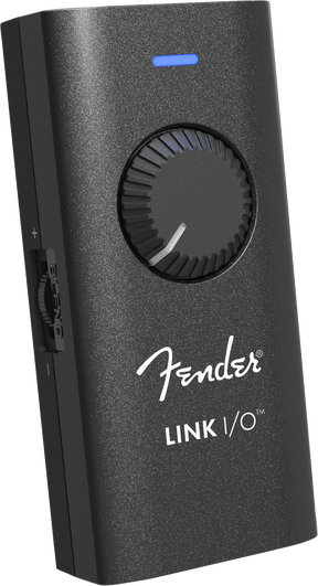 The image shows a Fender LINK I/O audio interface, a compact, portable device for connecting guitars or basses to computers, tablets, or smartphones. It features a black textured housing with the white Fender logo, a large central rotary knob, a blue LED indicator, and multiple side ports for inputs and outputs. Designed for recording, practice, and music production, it enables seamless integration with digital audio workstations and amp simulation software.
