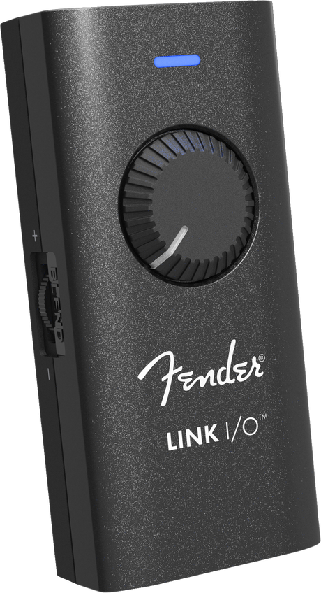 The image shows a Fender LINK I/O audio interface, a compact, portable device for connecting guitars or basses to computers, tablets, or smartphones. It features a black textured housing with the white Fender logo, a large central rotary knob, a blue LED indicator, and multiple side ports for inputs and outputs. Designed for recording, practice, and music production, it enables seamless integration with digital audio workstations and amp simulation software.