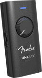 The image shows a Fender LINK I/O audio interface, a compact, portable device for connecting guitars or basses to computers, tablets, or smartphones. It features a black textured housing with the white Fender logo, a large central rotary knob, a blue LED indicator, and multiple side ports for inputs and outputs. Designed for recording, practice, and music production, it enables seamless integration with digital audio workstations and amp simulation software.