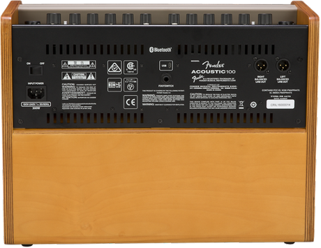 The image shows the back panel of an Orange Acoustic Crush 100 guitar amplifier. It features the signature orange/tan cabinet, black control panel, and model label. Visible are Bluetooth connectivity, multiple inputs/outputs including XLR and audio jacks, power connections, ventilation slots, safety certifications, and a wooden cabinet. This solid-state amp is designed for acoustic guitars and delivers 100 watts, suitable for live use.