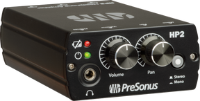 The image shows a PreSonus HP2 headphone amplifier, a compact black unit used in studios for headphone distribution. Visible features include a headphone jack, volume and pan knobs, stereo/mono switch, power and mute buttons, and red/green LED indicators. It allows multiple users to monitor audio with individual volume and stereo control by connecting to mixers or audio interfaces.