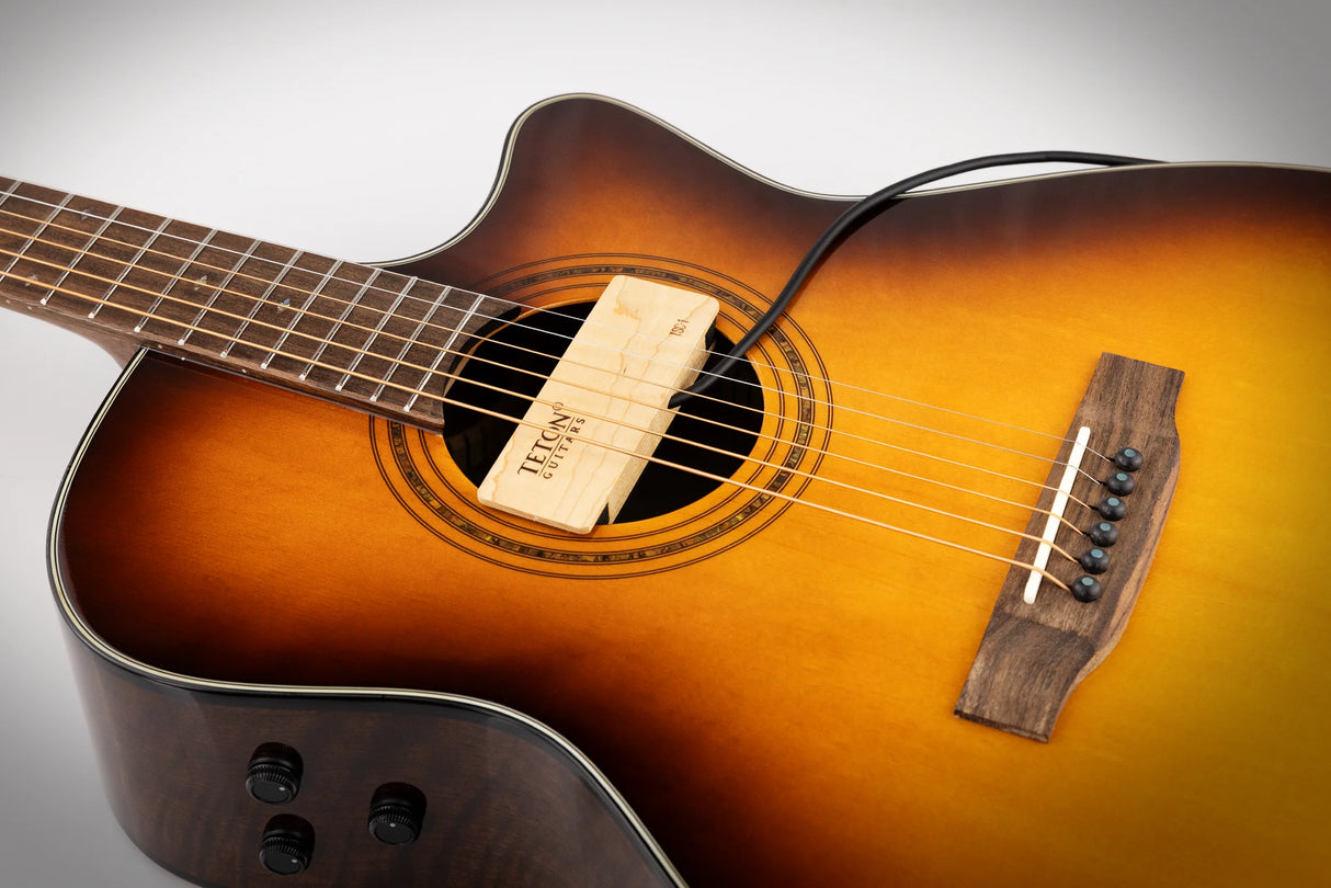 The image shows a beautiful acoustic-electric guitar with a sunburst finish, transitioning from dark edges to a golden center. It features a cutaway body for easy access to higher frets, a sound hole with a decorative rosette, a dark wooden bridge with black pins, a dark fretboard with markers, control knobs for amplification, and a wooden humidifier labeled "TEKNO GUITARS" inside the sound hole.