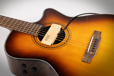 The image shows a beautiful acoustic-electric guitar with a sunburst finish, transitioning from dark edges to a golden center. It features a cutaway body for easy access to higher frets, a sound hole with a decorative rosette, a dark wooden bridge with black pins, a dark fretboard with markers, control knobs for amplification, and a wooden humidifier labeled "TEKNO GUITARS" inside the sound hole.