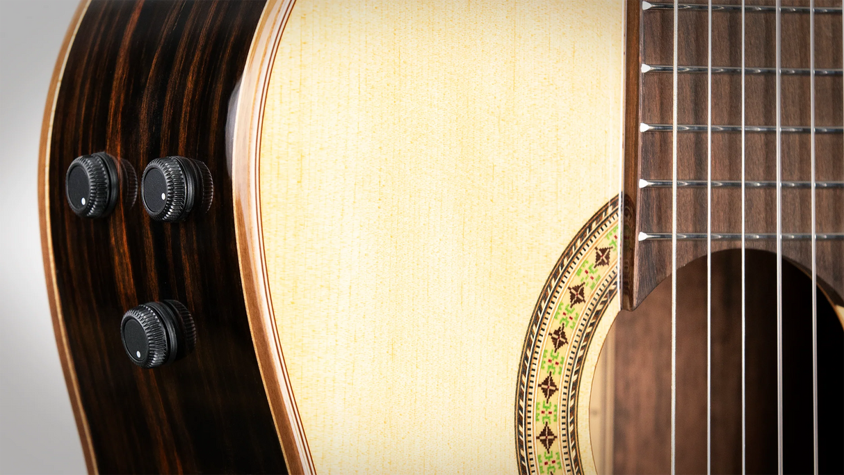 The image shows a close-up of an acoustic-electric guitar, featuring the sound hole, part of the fretboard with strings, and the body. Three black control knobs for adjusting the electric pickup’s volume and tone are visible on the side. The wood grain and rosette design around the sound hole are also clearly shown, highlighting the guitar’s acoustic and electric features.