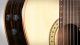 The image shows a close-up of an acoustic-electric guitar, featuring the sound hole, part of the fretboard with strings, and the body. Three black control knobs for adjusting the electric pickup’s volume and tone are visible on the side. The wood grain and rosette design around the sound hole are also clearly shown, highlighting the guitar’s acoustic and electric features.