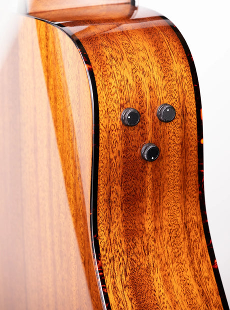 The image shows the side and back of an acoustic-electric guitar with rich, warm-toned wood and prominent grain, likely mahogany. It features three black control knobs for built-in electronics, dark binding along the edges, and a glossy, polished finish that emphasizes the wood’s natural beauty. This well-crafted guitar combines traditional acoustic design with modern amplification, indicating a mid to high-end instrument.