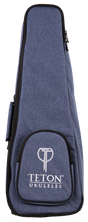 The image shows a blue denim-like ukulele gig bag with the "TETON UKULELES" logo on the front pocket. It features a zippered closure around the perimeter, a front storage pocket, and carrying straps (partially visible). This padded soft case is designed to protect and transport a ukulele, offering a lightweight, protective option. Teton is a well-known brand for acoustic instruments and accessories.
