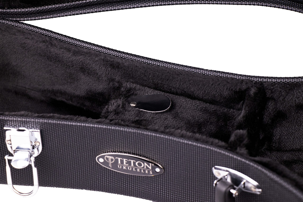 The image shows the interior of a Teton Ukuleles hard case, featuring a plush black velvet lining molded to fit a ukulele. It has a textured black exterior, chrome latches and hinges, and a clear "TETON UKULELES" logo inside. This professional-grade case offers durable protection and a sleek design, reflecting Teton’s commitment to quality instrument accessories.