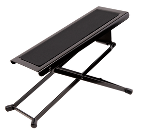 The image shows a guitar footstool with a black metal X-shaped folding frame, a padded cushioned top, adjustable height, and rubber feet for stability. It’s designed for classical and acoustic guitarists to elevate the left foot, promoting proper posture and better control. Portable and foldable, it’s a common accessory in music schools, concert halls, and practice rooms.