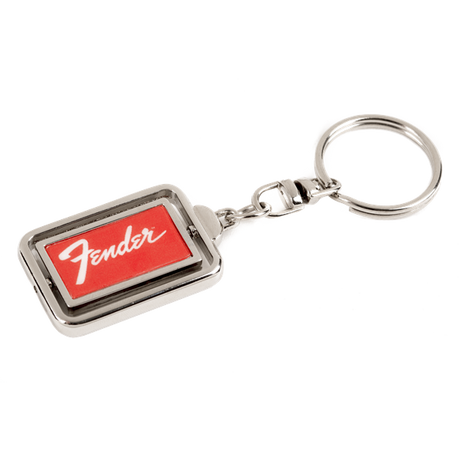 The image shows a metal Fender keychain with a rectangular design featuring the red Fender logo on a dark background, framed by chrome-colored metal. It includes a standard key ring attached by a small chain link. This branded accessory represents Fender, a renowned guitar and amplifier manufacturer popular among musicians and fans.