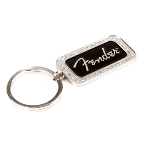The image shows a Fender keychain featuring a rectangular metal charm with a black center displaying the white Fender logo in signature script. The charm has a decorative beaded border and is attached to a standard split-ring. This music-themed accessory is popular among musicians and fans as a collectible or gift, representing the iconic brand known for electric guitars and amplifiers.