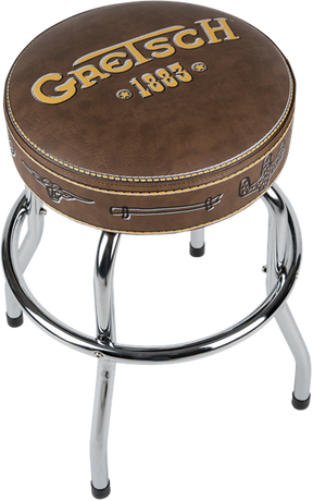 The image shows a Gretsch-branded bar stool with a round brown leather seat featuring the "Gretsch" logo and "1883" in gold lettering. It has chrome metal legs, decorative stitching, and small Gretsch logo details on the seat edges. This music-themed stool is branded merchandise designed for musicians or fans, typically used in studios or performance spaces to showcase the iconic Gretsch brand.