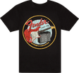 The image shows a black t-shirt with a vintage-style circular Fender logo. It features "Fender" in red script, "Since 1946," a red Stratocaster-style guitar, and a guitar amplifier. The bottom of the circle reads "GUITARS & AMPLIFIERS." This design highlights Fender's legacy as a leading maker of electric guitars and amplifiers.