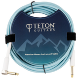 The image shows a Teton Guitars Premium Woven Instrument Cable with a light blue/turquoise woven jacket for durability and style. It has one straight and one right-angle 1/4" (6.35mm) gold-plated plug. The cable is coiled and secured with velcro, displayed in branded packaging featuring a mountain-themed design. This cable is ideal for connecting guitars, basses, and other instruments to audio gear, offering enhanced protection and signal quality.