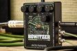 The image shows the Electro-Harmonix 15 Watt Howitzer, a compact power amp guitar pedal. It features four top knobs labeled VOL, TREBLE, MID, and BASS, a GAIN knob on the right, and a BRIGHT switch. The green and black pedal has military-themed graphics, including a howitzer illustration. Input/output jacks are on the sides, and it requires 24VDC at 1.0A. Designed to add amp power and tone shaping to a guitar setup.