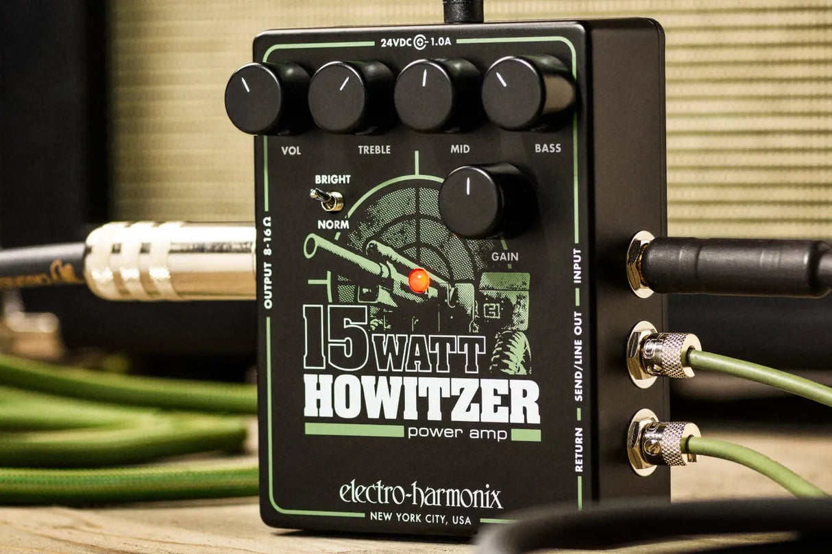 The image shows the Electro-Harmonix 15 Watt Howitzer, a compact power amp guitar pedal. It features four top knobs labeled VOL, TREBLE, MID, and BASS, a GAIN knob on the right, and a BRIGHT switch. The green and black pedal has military-themed graphics, including a howitzer illustration. Input/output jacks are on the sides, and it requires 24VDC at 1.0A. Designed to add amp power and tone shaping to a guitar setup.