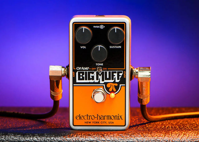 The image shows an Electro-Harmonix Big Muff guitar effects pedal with a classic orange and black design. It features Volume, Sustain, and Tone knobs, an Op Amp switch, a 9V power input, and a true bypass footswitch. Two 1/4" cables are connected. Marked "New York City, USA," this compact pedal delivers the iconic thick, creamy fuzz tone known for rich, wall-of-sound guitar effects.
