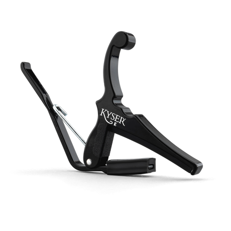 The image shows a black Kyser guitar capo with a spring-loaded mechanism for quick, one-handed use. It features a padded contact surface to protect the guitar neck, an ergonomic handle for easy grip, and the "KYSER" brand name visible. This popular capo lets guitarists quickly change keys without retuning, ideal for acoustic players and singer-songwriters.