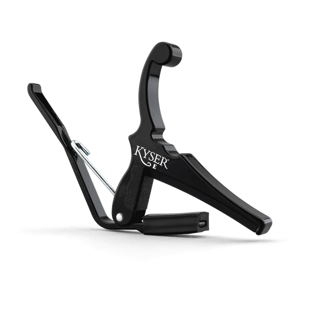 The image shows a black Kyser guitar capo with a spring-loaded mechanism for quick, one-handed use. It features a padded contact surface to protect the guitar neck, an ergonomic handle for easy grip, and the "KYSER" brand name visible. This popular capo lets guitarists quickly change keys without retuning, ideal for acoustic players and singer-songwriters.