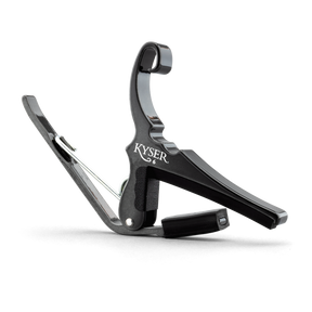 The image shows a Kyser model 6 guitar capo with a black finish. It features a spring-loaded, curved clamp design, an ergonomic handle for quick use, and a fabric strap for secure attachment. This popular quick-change capo is known for reliable clamping pressure and ease of use, favored by acoustic and electric guitar players. The capo is photographed against a white background.