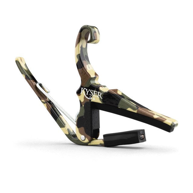 The image shows a KYSER spring-loaded guitar capo with a camouflage pattern in green, brown, and tan. It features a curved metal frame, a padded bar to press the strings, and a strap attachment point. This quick, one-handed capo evenly clamps all strings for clean sound and is popular among guitarists for easy use during performances.