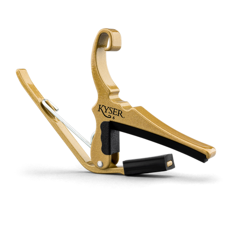 The image shows a Kyser brand guitar capo with a gold/brass metal frame and black padded bar. It features a curved hook shape, spring-loaded mechanism for one-handed use, and black padding to protect the guitar neck. Capos clamp all strings at a fret to raise pitch. Kyser capos are popular for their quick-change design and reliable grip.