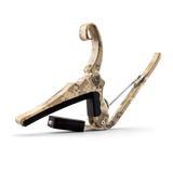 The image shows a guitar capo with a curved wooden frame featuring a tan and brown digital desert camouflage pattern. It has a black padded bar for pressing strings, a spring-loaded mechanism for easy attachment, metal hardware, and a visible logo on the frame. This capo allows guitarists to change string pitch by clamping the fretboard, offering a distinctive style compared to typical plain capos.