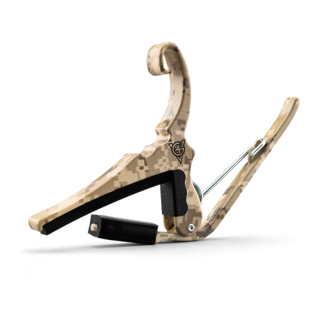 The image shows a guitar capo with a curved wooden frame featuring a tan and brown digital desert camouflage pattern. It has a black padded bar for pressing strings, a spring-loaded mechanism for easy attachment, metal hardware, and a visible logo on the frame. This capo allows guitarists to change string pitch by clamping the fretboard, offering a distinctive style compared to typical plain capos.