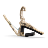 The image shows a Kyser Quick-Change guitar capo with a camouflage or wood-grain finish. It features a spring-loaded clamp with black padding to protect the fretboard and neck. The "KYSER" brand name is visible, and a black strap is attached for easy carrying. This popular accessory allows guitarists to quickly change pitch by clamping across the fretboard, enabling quick transposition with one-handed operation.