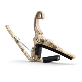 The image shows a Kyser Quick-Change guitar capo with a camouflage or wood-grain finish. It features a spring-loaded clamp with black padding to protect the fretboard and neck. The "KYSER" brand name is visible, and a black strap is attached for easy carrying. This popular accessory allows guitarists to quickly change pitch by clamping across the fretboard, enabling quick transposition with one-handed operation.