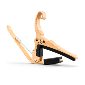 The image shows a Kyser guitar capo with a natural wooden finish. It features a spring-loaded, curved handle for one-handed use and black rubber padding to protect the fretboard and strings. The "KYSER" logo is visible, highlighting its brand. This ergonomic capo raises the pitch of all strings simultaneously, allowing guitarists to change keys easily while playing.