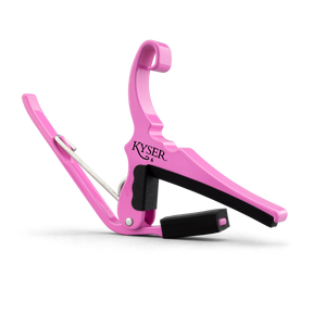 The image shows a bright pink Kyser guitar capo, a popular accessory that clamps across the fretboard to raise pitch and change keys easily. It features a spring-loaded mechanism for one-handed use, padded black contact surfaces to protect the guitar, and a curved design to fit the neck. Known for quick changes, Kyser capos are favored by guitarists seeking durability and convenience with a touch of personality.