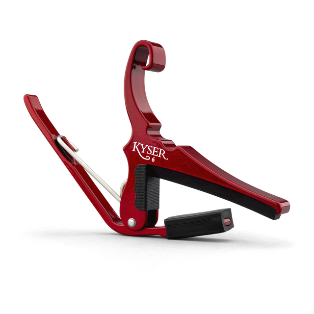The image shows a Kyser spring-loaded, trigger-style guitar capo with a red anodized aluminum finish. It has black rubber padding to protect the guitar strings and neck while providing a secure grip. This popular accessory allows quick key changes by clamping all strings at any fret. Its curved design fits the guitar neck for even pressure, and the spring mechanism enables easy one-handed use.