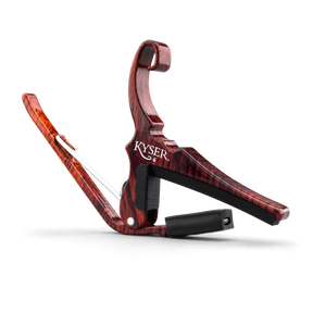The image shows a Kyser guitar capo with a reddish-brown wood grain finish resembling rosewood. It features a spring-loaded clamp and curved handle for one-handed use, a black rubber pad that presses the strings, and a black fabric strap for secure attachment. This quick-change style capo raises the pitch by clamping the fretboard, allowing guitarists to easily change keys while using familiar chord shapes.