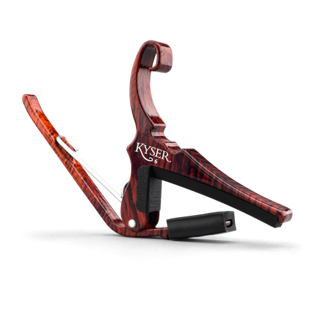 The image shows a Kyser guitar capo with a reddish-brown wood grain finish resembling rosewood. It features a spring-loaded clamp and curved handle for one-handed use, a black rubber pad that presses the strings, and a black fabric strap for secure attachment. This quick-change style capo raises the pitch by clamping the fretboard, allowing guitarists to easily change keys while using familiar chord shapes.