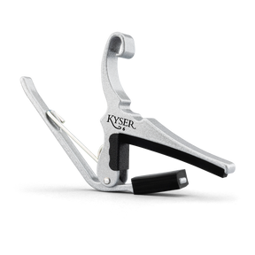 The image shows a KYSER guitar capo made of silver metal with black rubber padding. It features a spring-loaded clamp with a curved handle for easy one-handed use. The capo clamps across the guitar fretboard to raise the pitch of all strings simultaneously, allowing players to change keys with the same chord shapes. KYSER is a trusted brand known for reliable, quick-change capos.