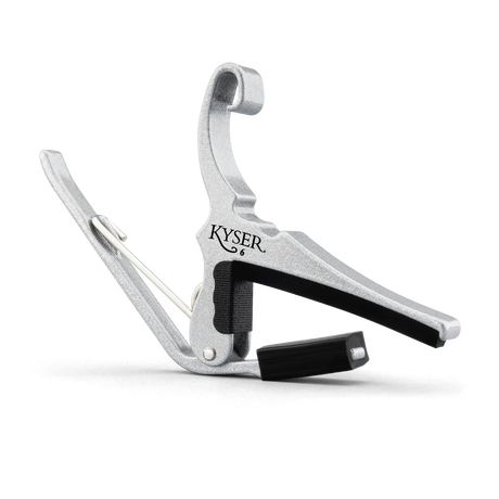 The image shows a KYSER guitar capo made of silver metal with black rubber padding. It features a spring-loaded clamp with a curved handle for easy one-handed use. The capo clamps across the guitar fretboard to raise the pitch of all strings simultaneously, allowing players to change keys with the same chord shapes. KYSER is a trusted brand known for reliable, quick-change capos.