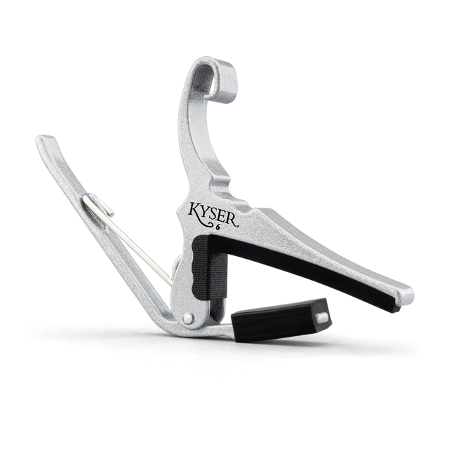 The image shows a KYSER guitar capo made of silver metal with black rubber padding. It features a spring-loaded clamp with a curved handle for easy one-handed use. The capo clamps across the guitar fretboard to raise the pitch of all strings simultaneously, allowing players to change keys with the same chord shapes. KYSER is a trusted brand known for reliable, quick-change capos.