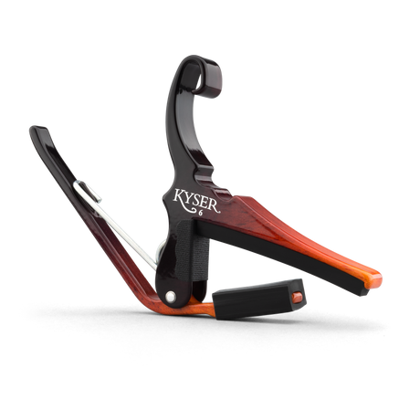 The image shows a Kyser guitar capo with a wood-grain finish in reddish-brown tones. This spring-loaded clamp attaches to the fretboard, raising the pitch of all strings for easy key changes without new chords. It features padded surfaces to protect the guitar and a quick-release handle for fast adjustments. Kyser capos are trusted by acoustic and electric guitarists for reliability and ease of use.
