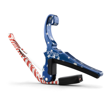 The image shows a Kyser spring-loaded guitar capo with a patriotic American flag design, featuring blue with white stars and red and white stripes. This essential accessory clamps the fretboard to raise string pitch, allowing easy key changes. Kyser capos are popular among guitarists for their functionality and quick one-handed use, combining practicality with a distinctive, decorative look.