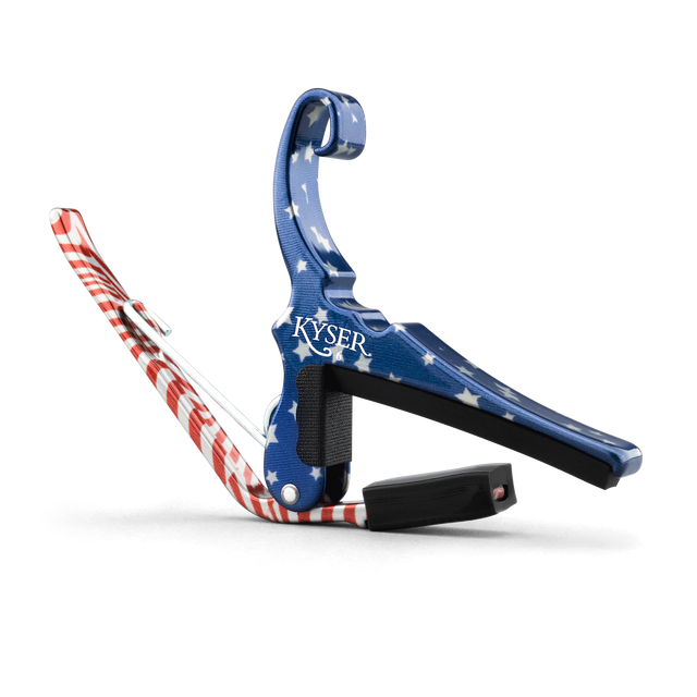 The image shows a Kyser spring-loaded guitar capo with a patriotic American flag design, featuring blue with white stars and red and white stripes. This essential accessory clamps the fretboard to raise string pitch, allowing easy key changes. Kyser capos are popular among guitarists for their functionality and quick one-handed use, combining practicality with a distinctive, decorative look.