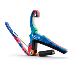 The image shows a colorful Kyser guitar capo with a vibrant tie-dye pattern featuring blues, reds, and oranges. It is spring-loaded with a curved clamp and has black padding to protect the guitar neck and strings. A small black cylindrical pick holder is integrated into the design. This accessory clamps the fretboard to change string pitch, combining functionality with a bright, artistic look.