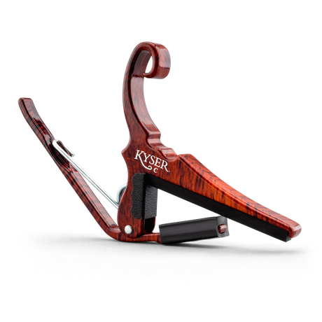 The image shows a KYSER spring-loaded guitar capo with a reddish-brown wood grain finish. It features a curved clamp arm, a padded black bar to protect strings, and a quick-release handle for easy one-handed use. Designed to clamp the fretboard and raise string pitch, this elegant capo is a practical and stylish accessory that helps guitarists change keys and achieve different chord voicings effortlessly.
