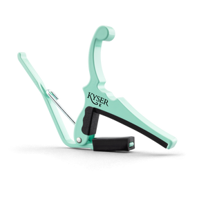The image shows a Kyser guitar capo in a mint green finish. This spring-loaded capo has a curved metal frame, black rubber padding to protect the strings and fretboard, and an ergonomic handle for easy one-handed use. Kyser is known for quick-change capos popular with acoustic and electric guitarists for fast fret adjustments during performances.