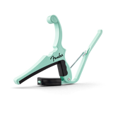 The image shows a mint green Fender guitar capo with the black Fender logo in script. It features a spring-loaded mechanism, a black rubber-padded clamp to press the strings, and a curved arm to open and position it on the guitar neck. This accessory changes string pitch by shortening their length, enabling different keys with the same chord shapes. Its clean design combines functionality and style.