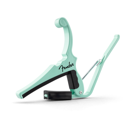 The image shows a mint green Fender guitar capo with the black Fender logo in script. It features a spring-loaded mechanism, a black rubber-padded clamp to press the strings, and a curved arm to open and position it on the guitar neck. This accessory changes string pitch by shortening their length, enabling different keys with the same chord shapes. Its clean design combines functionality and style.