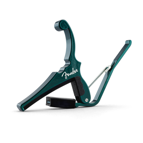 The image shows a teal Fender spring-loaded trigger capo with a white Fender logo. It features a padded black bar that clamps across the guitar fretboard and a curved handle for easy one-handed use. This sleek capo allows guitarists to change keys quickly while maintaining familiar chord shapes, combining functionality with Fender’s classic design, and is popular for acoustic playing.