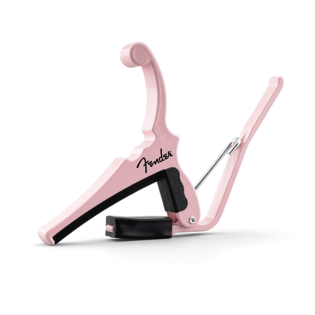 The image shows a Fender guitar capo in a stylish pink/shell pink color. It features a spring-loaded, curved clamp design with black padding to protect the guitar neck. Made of durable metal with a smooth finish, it has an ergonomic handle for easy use. This reliable capo evenly presses all strings, helping guitarists change keys while maintaining tuning and chord fingerings.