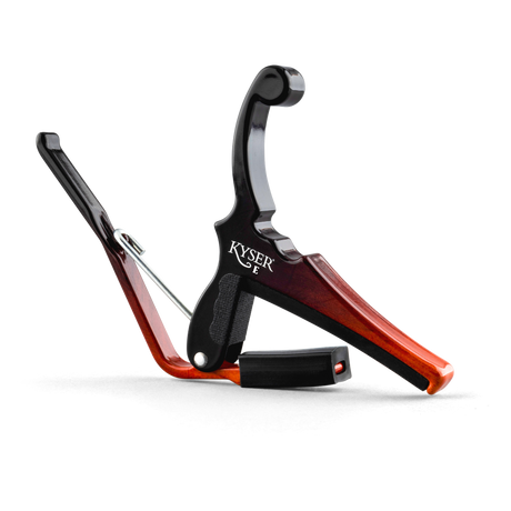 The image shows a Kyser guitar capo with a spring-loaded black metal clamp and a reddish-brown wooden finish. It features an ergonomic handle for one-handed use and black padded surfaces to protect the guitar neck and strings. Designed to clamp across the fretboard, it raises the pitch of all strings simultaneously, allowing quick key changes without retuning, making it popular for acoustic and electric guitars.